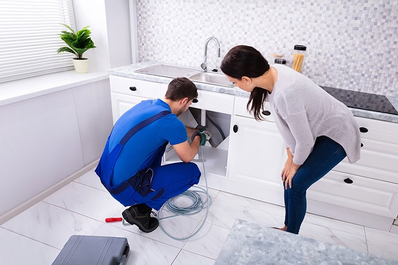 Why You Should Never Ignore a Slow Kitchen Sink Drain. Photo of a woman with a plumber working on her kitchen sink drain.