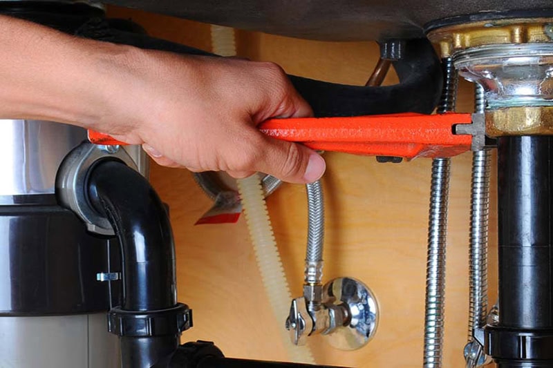 2025 Guide to Garbage Disposals. Photo of a plumber's hand performing maintenance on a garbage disposal under a kitchen sink.